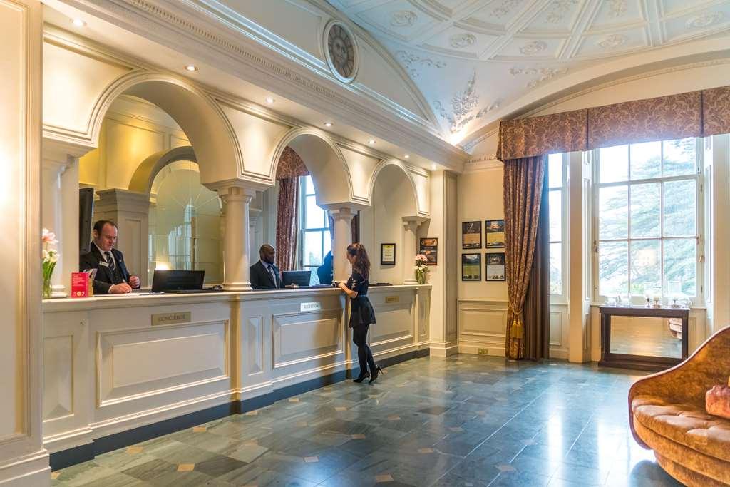 Macdonald Bath Spa Hotel Interieur foto The photo shows the interior of a hotel reception area. There is a stylish front desk with several staff members attending to guests. A woman is interacting with one of the receptionists. The design features elegant architectural details, including a