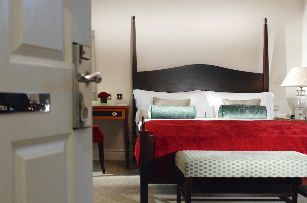 Macdonald Bath Spa Hotel Kamer foto The photo shows a well-decorated hotel room. In the foreground, there is a partially opened door leading into the room. The focal point of the room is a large bed with a dark wooden headboard and stylish red and white bedding, accented with green pil