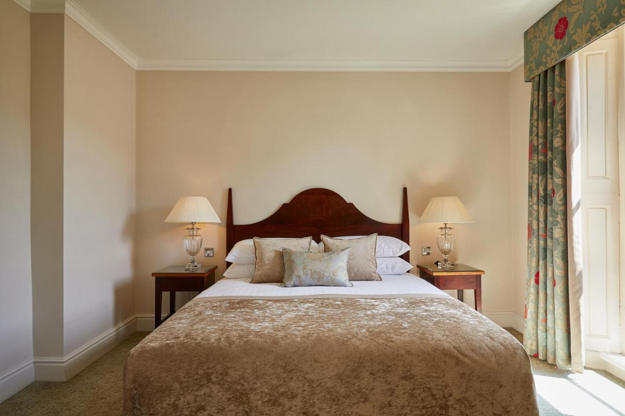 Macdonald Bath Spa Hotel Buitenkant foto The photo shows a cozy hotel bedroom. It features a large bed with a plush, light-colored bedspread and several pillows. On either side of the bed, there are wooden nightstands with lamps providing soft lighting. The walls are painted a neutral color