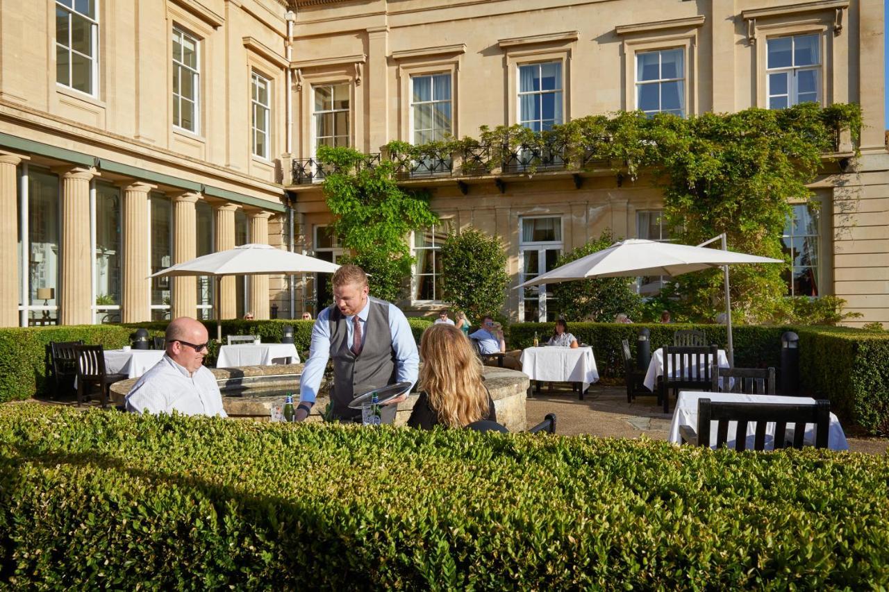 Macdonald Bath Spa Hotel Buitenkant foto The photo depicts an outdoor dining area of a restaurant. In the foreground, there are neatly trimmed hedges surrounding the dining space. Two patrons are seated at a table; one is a man wearing sunglasses and the other is a woman facing him. A waite
