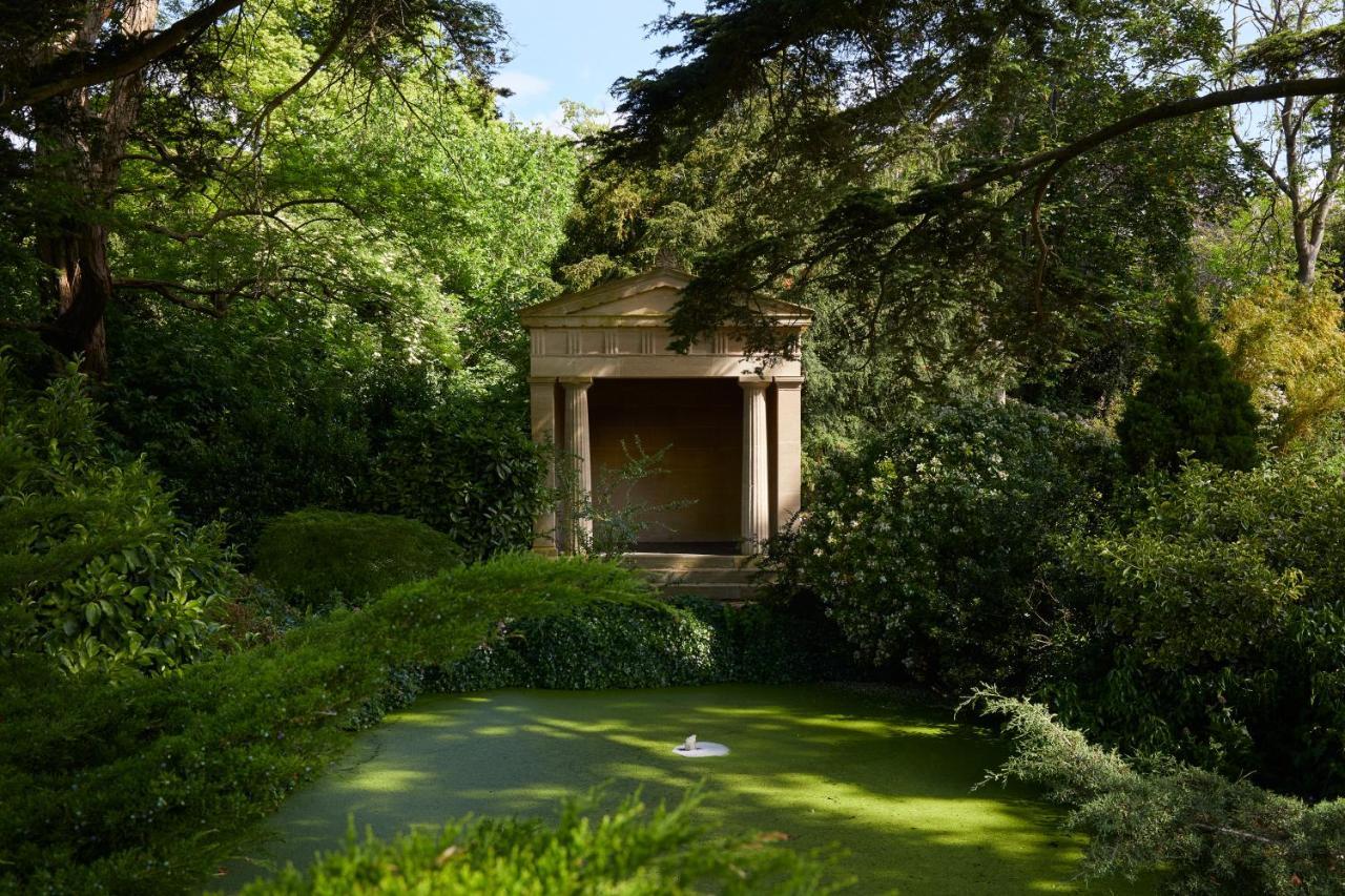 Macdonald Bath Spa Hotel Buitenkant foto The photo shows a serene garden scene featuring lush greenery and a tranquil pond. In the background, there is a classical-looking structure with columns, resembling a small temple or pavilion. The pond appears to have vegetation covering its surface
