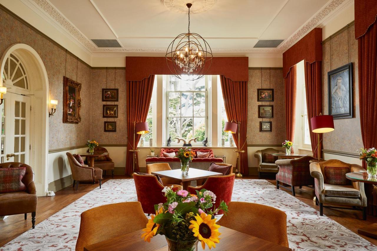 Macdonald Bath Spa Hotel Buitenkant foto The photo shows a well-decorated living room or lounge area. The space features elegant furnishings, including chairs and a round table at the center. Large windows allow natural light to fill the room, draped with rich red curtains. The walls are ad