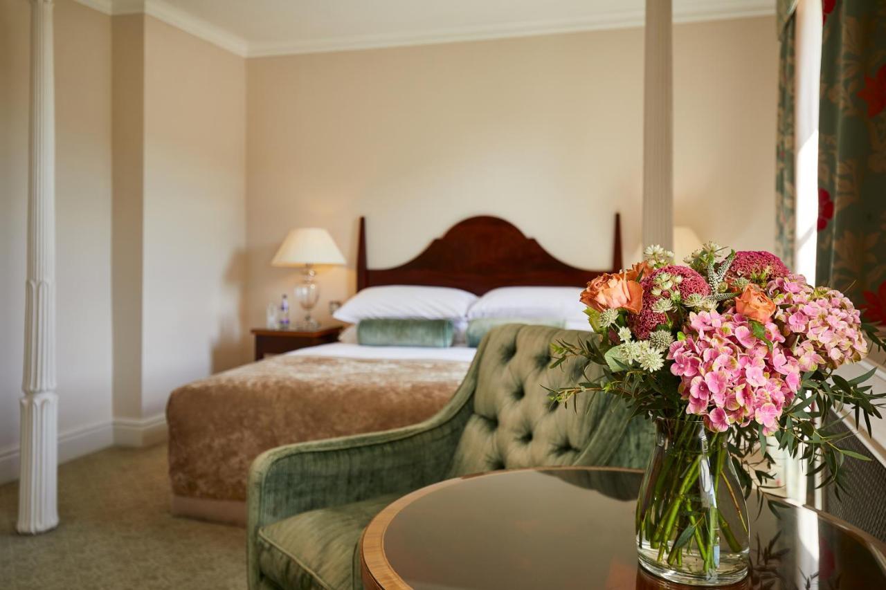 Macdonald Bath Spa Hotel Buitenkant foto The photo shows a cozy bedroom featuring a large bed with a wooden headboard, topped with a fluffy blanket. To one side, there is a small bedside lamp providing soft lighting. In the foreground, there is an elegant green chair with a tufted design an