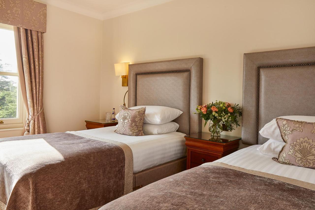 Macdonald Bath Spa Hotel Buitenkant foto The photo shows a hotel room featuring two beds with elegant headboards. Each bed is neatly made with white linens and a soft, decorative throw blanket. There are matching pillows on the beds, and a small, stylish bouquet of flowers is placed on a wo
