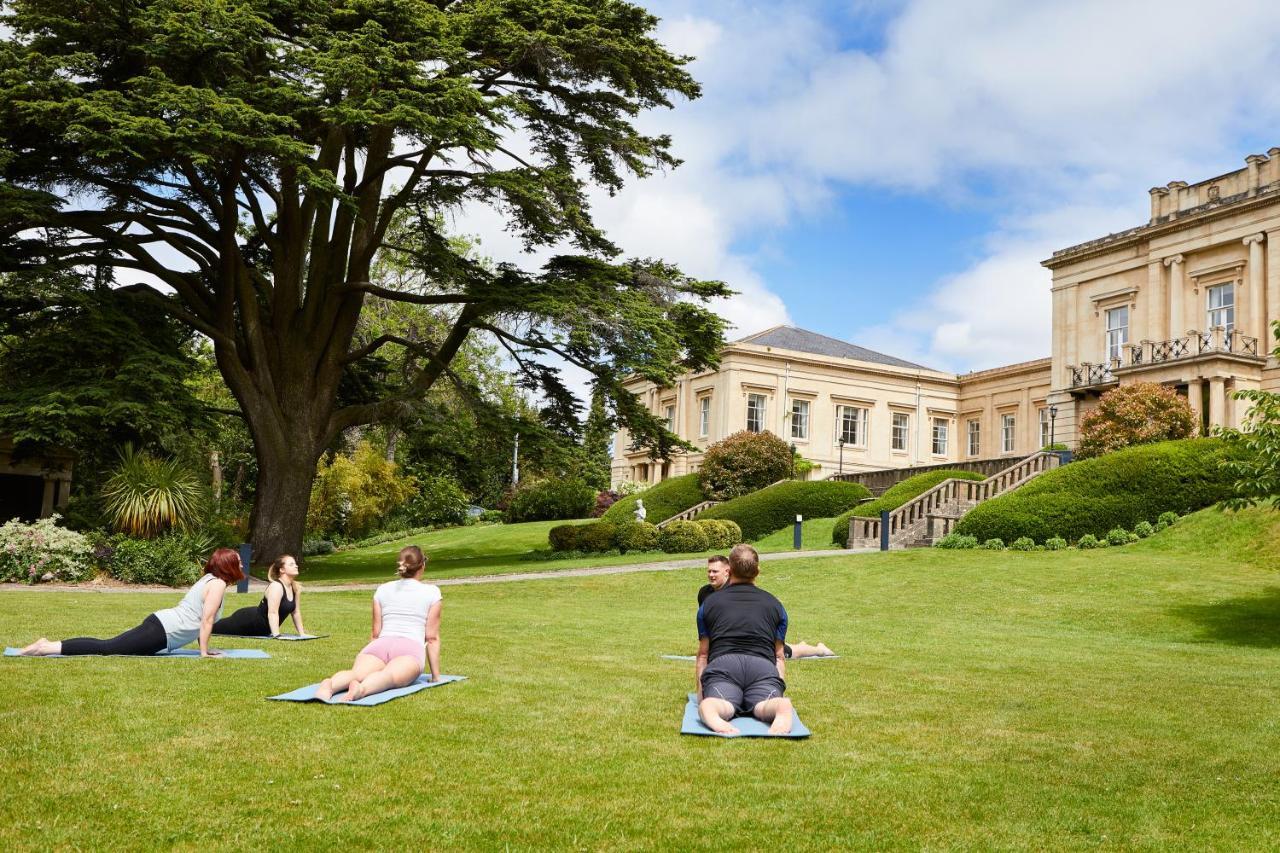 Macdonald Bath Spa Hotel Buitenkant foto The photo shows a group of people engaging in a yoga session outdoors. They are positioned on yoga mats on a lush green lawn in front of a large, elegant building with columns. Behind them, there are trees and well-maintained gardens, contributing to