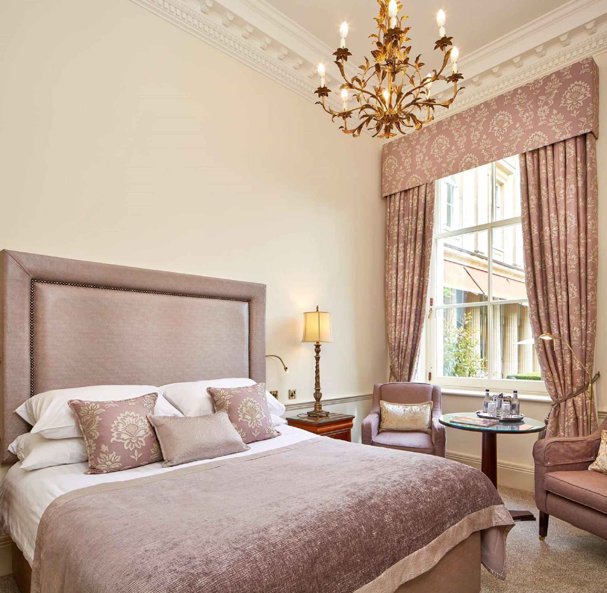 Macdonald Bath Spa Hotel Buitenkant foto The photo shows a beautifully decorated hotel room. It features a large bed with a plush headboard, adorned with several decorative pillows. The walls are painted in a soft color, and there is a chandelier hanging from the ceiling. Natural light come