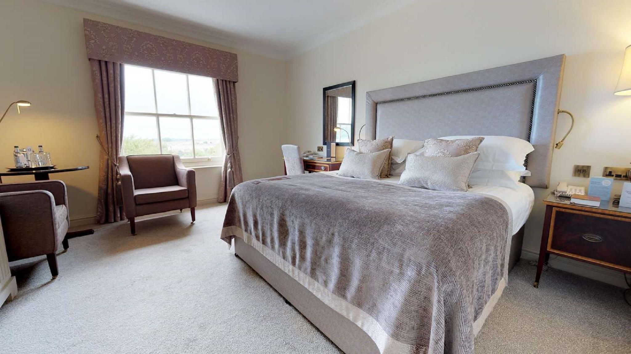 Macdonald Bath Spa Hotel Buitenkant foto The photo shows a well-furnished bedroom. It features a large bed with a light gray headboard and several decorative pillows on top. The bed is covered with a soft gray blanket. To one side of the room, there is a comfortable armchair and a small tab