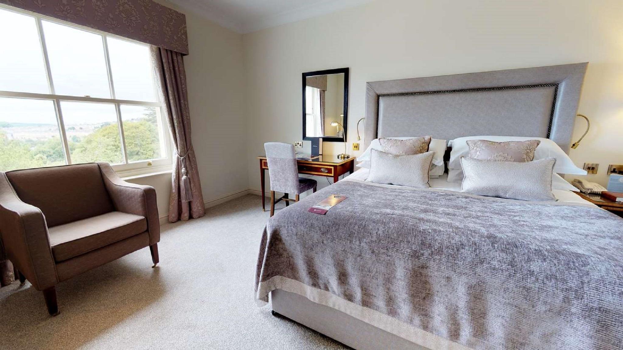 Macdonald Bath Spa Hotel Buitenkant foto The photo shows a cozy and well-decorated bedroom. It features a large bed with a plush, grey bedspread and several decorative pillows. Beside the bed, there is a wooden bedside table with a lamp. A small desk and chair are positioned near a window, 