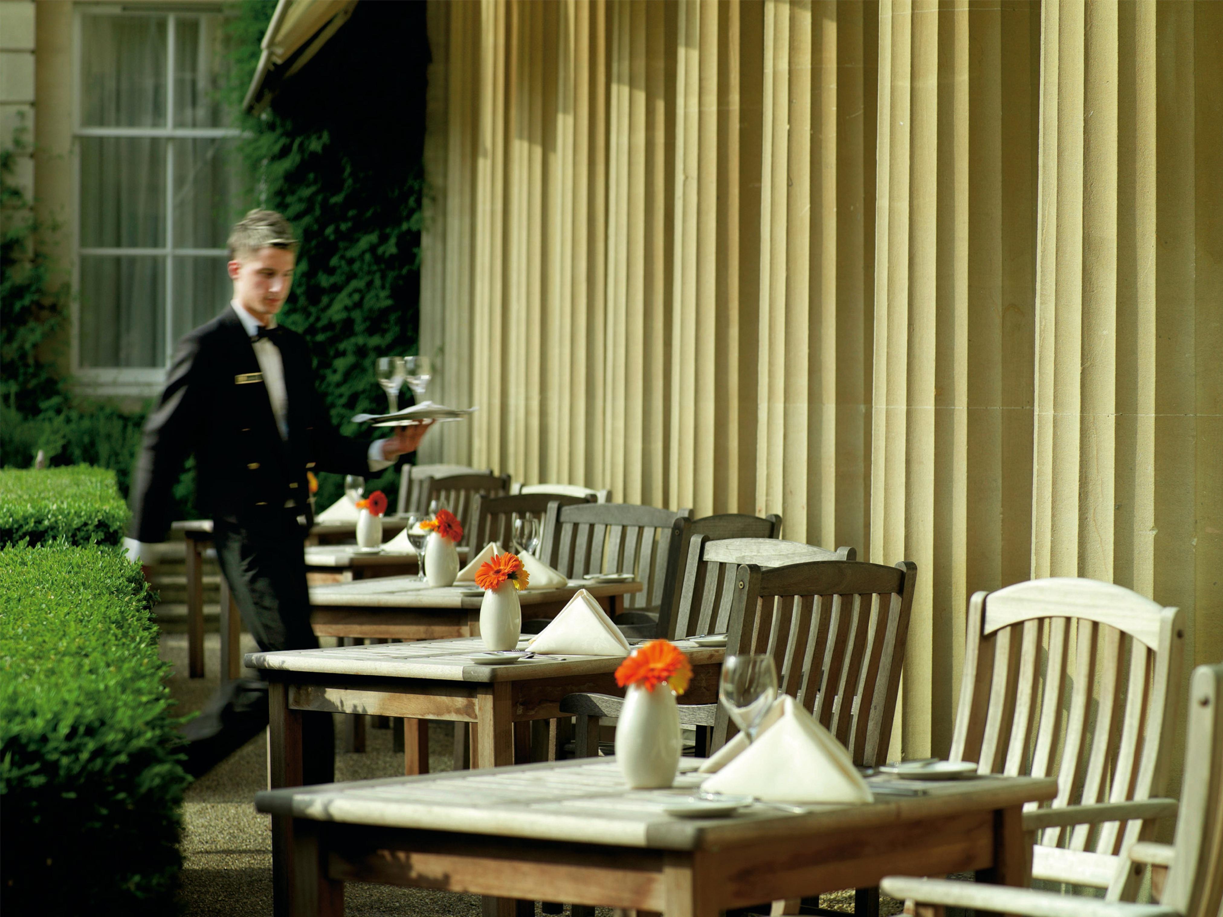 Macdonald Bath Spa Hotel Buitenkant foto The photo shows an outdoor dining area, featuring several wooden tables and chairs arranged neatly. Each table is set with a white tablecloth and small vases containing flowers, adding a vibrant touch. A waiter in a formal black suit is seen moving t
