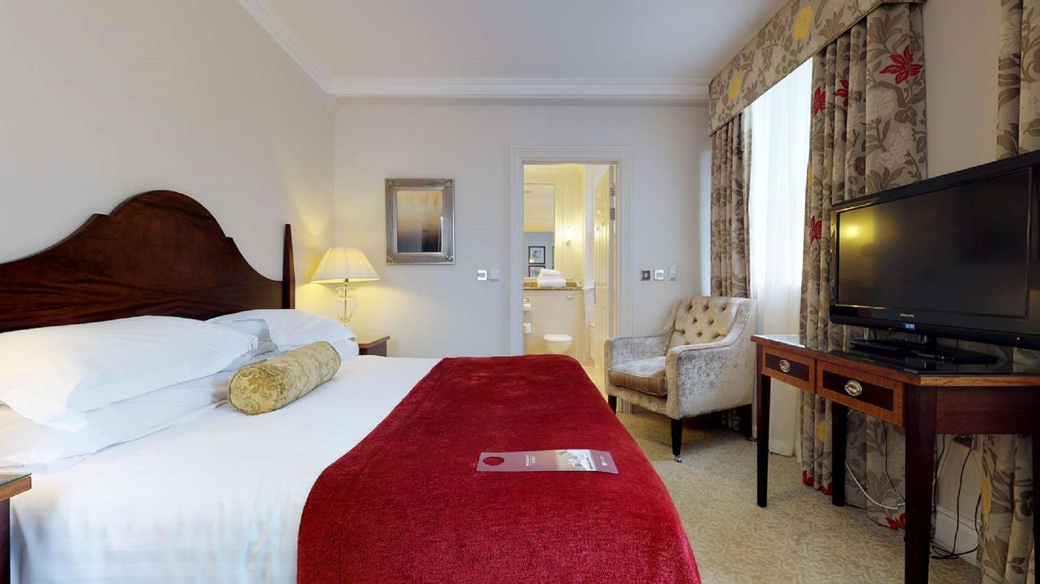 Macdonald Bath Spa Hotel Buitenkant foto The photo shows a hotel room with a queen-sized bed covered in white linens and a deep red throw blanket. There is a decorative pillow on the bed. Next to the bed, there's a small nightstand with a lamp. In the background, a seating area features a b