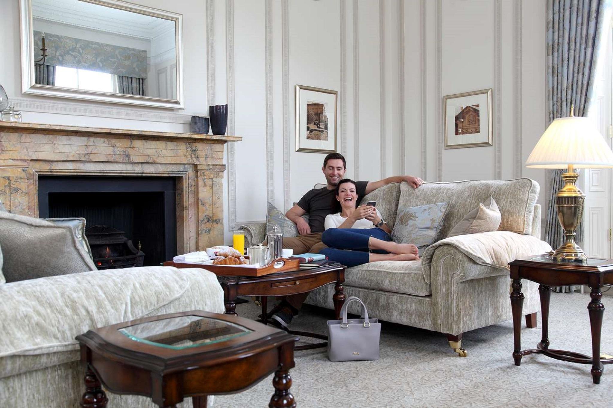 Macdonald Bath Spa Hotel Buitenkant foto The photo shows a cozy living room setting where a couple is sitting together on a light-colored sofa. They appear relaxed, with the man sitting slightly behind the woman, who is smiling and looking at her phone. The room features elegant decor, incl