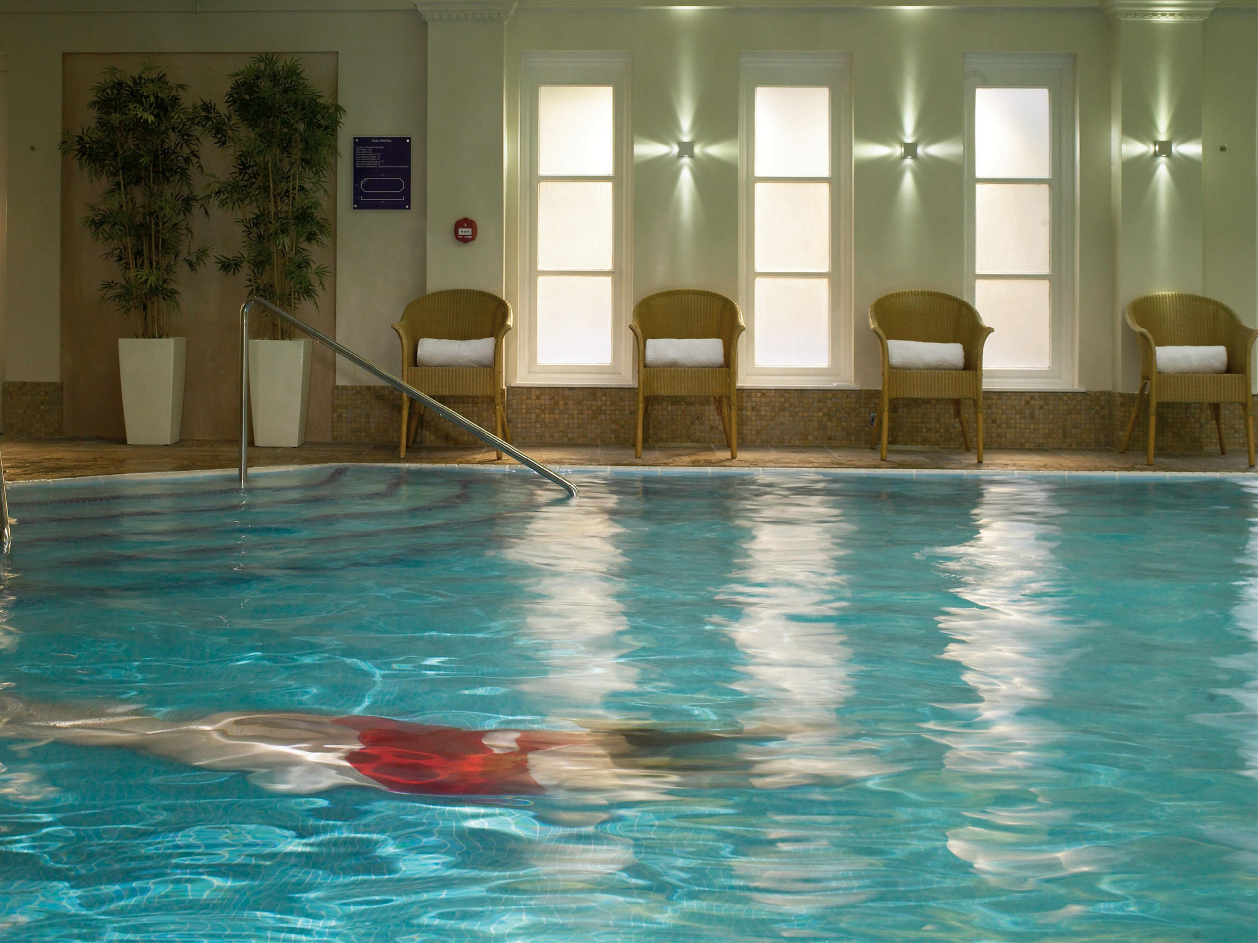 Macdonald Bath Spa Hotel Buitenkant foto The photo shows an indoor swimming pool with clear, blue water. In the pool, a person is swimming, partially submerged, and wearing a red swimsuit. Surrounding the pool, there are several empty chairs arranged along the side, along with potted plants