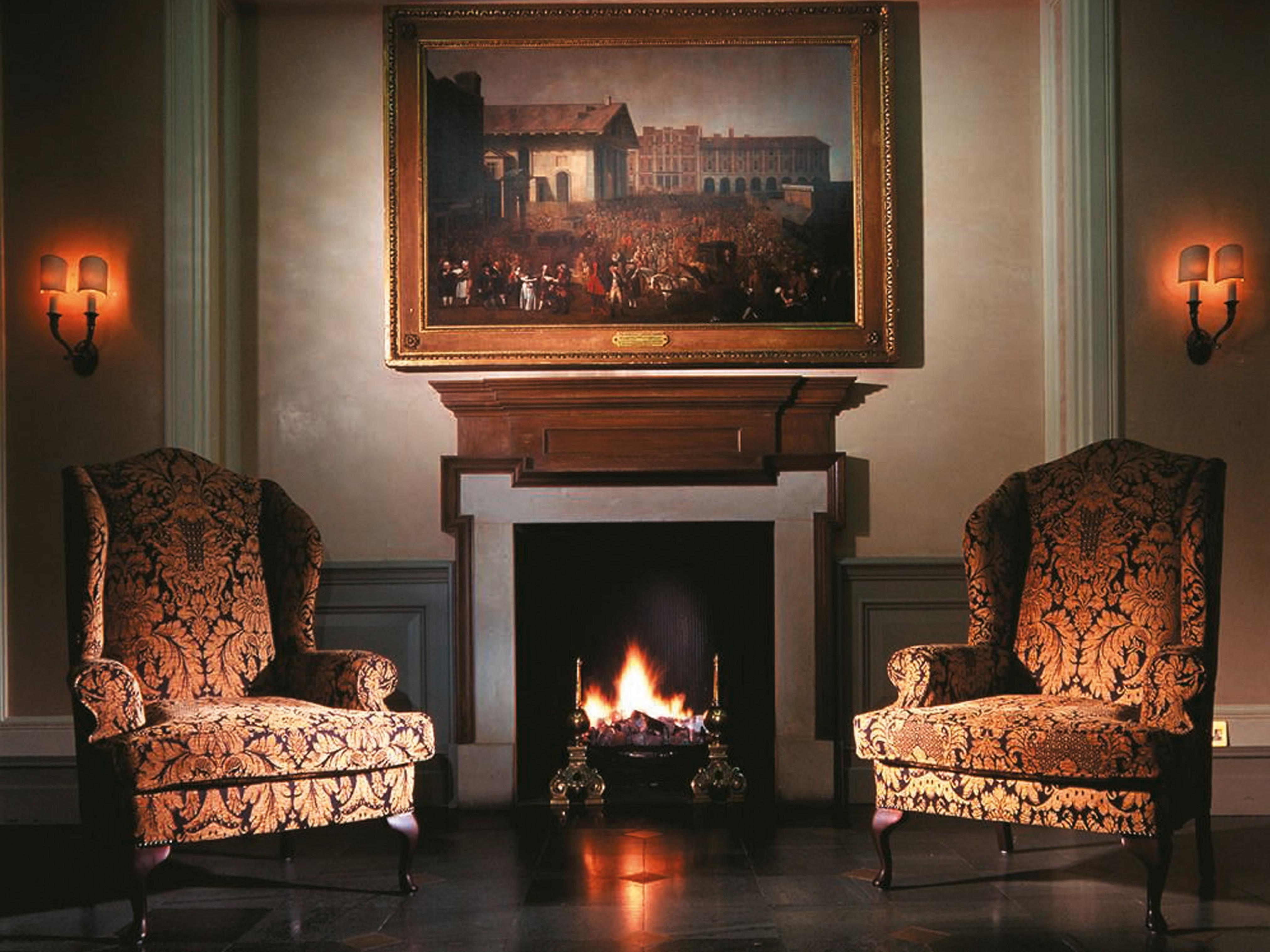 Macdonald Bath Spa Hotel Buitenkant foto The image shows a cozy indoor setting featuring two ornate armchairs placed opposite a fireplace. Above the fireplace hangs a large painting in a decorative frame. The warm glow from the fire illuminates the room, and there are wall sconces providing