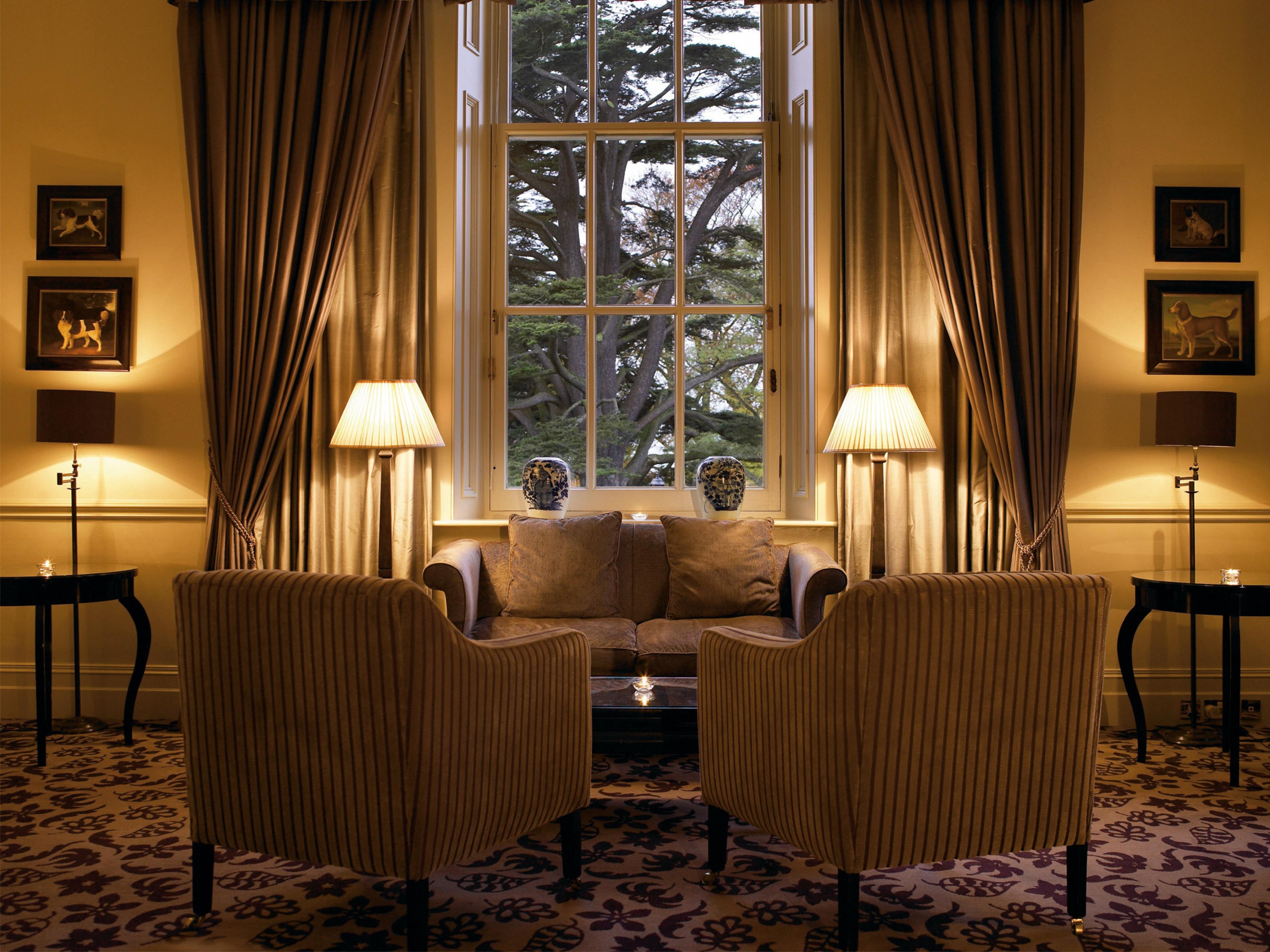 Macdonald Bath Spa Hotel Buitenkant foto The photo shows a cozy and elegant sitting area. There are two upholstered armchairs facing a small sofa, all arranged around a central coffee table. Soft lighting is provided by two lamps with white shades, one beside each chair. The window behind t