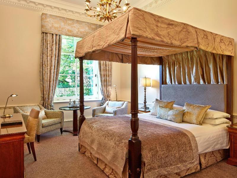 Macdonald Bath Spa Hotel Kamer foto The photo shows an elegantly designed bedroom featuring a four-poster bed with a decorative canopy. The bed is dressed with luxurious bedding and pillows, creating a cozy and inviting atmosphere. Flanking the bed are two comfortable armchairs and a s