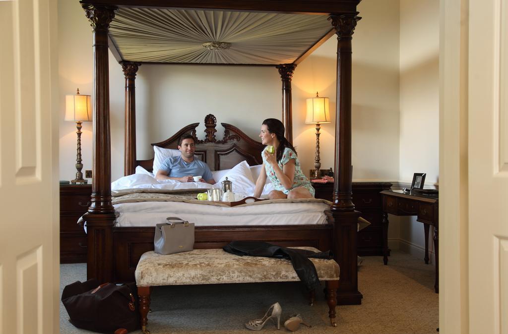Macdonald Bath Spa Hotel Buitenkant foto The image depicts a spacious, well-lit bedroom featuring a large four-poster bed with an ornate headboard. A man and a woman are sitting on the bed, appearing to engage in conversation. The woman is dressed in a patterned outfit, while the man is wea