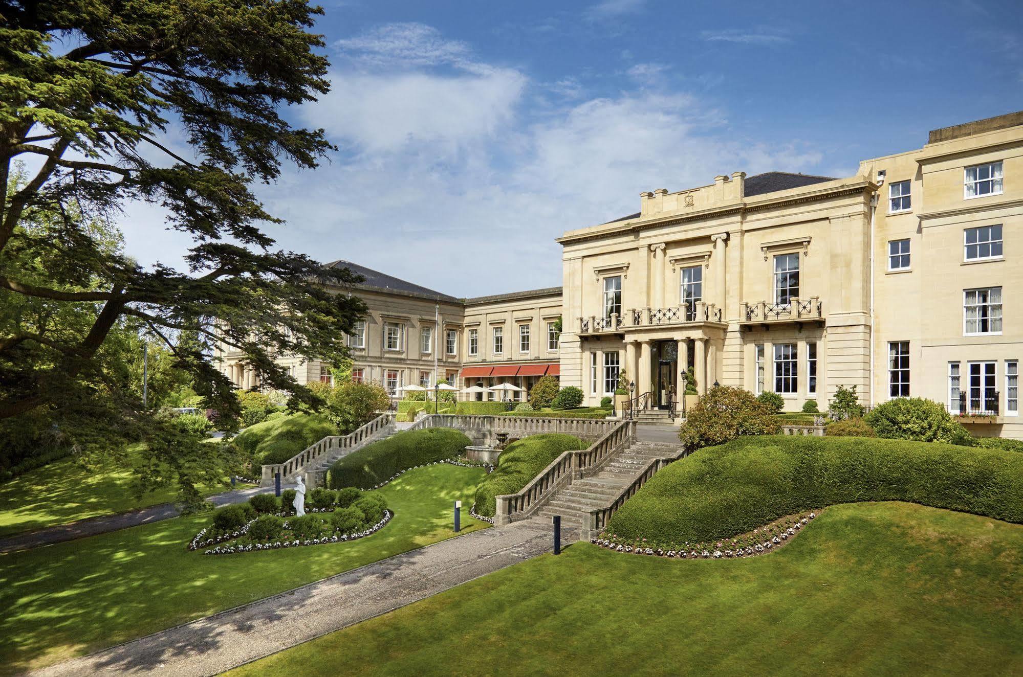 Macdonald Bath Spa Hotel Buitenkant foto The photo shows a grand building, likely a hotel or estate, surrounded by well-manicured gardens and lawns. The architecture appears elegant, with multiple stories and large windows. There are steps leading up to the entrance, and the setting include