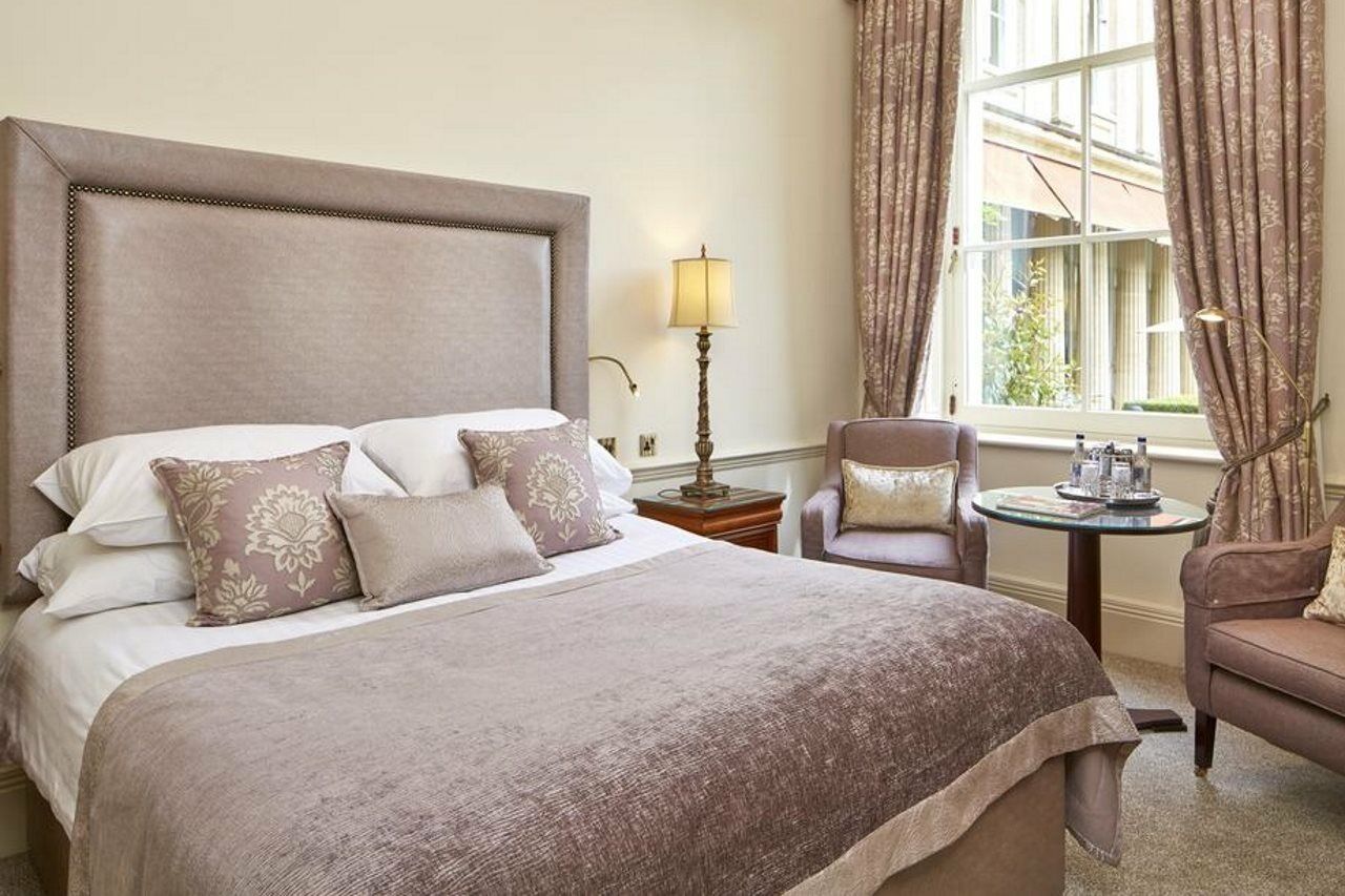 Macdonald Bath Spa Hotel Buitenkant foto The photo shows a stylish and elegantly decorated hotel room. It features a large bed with soft linens and several decorative pillows. The bedspread is a muted shade of beige or taupe. Beside the bed, there's a small table with two armchairs. One of 